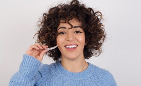 Woman with curly hair smiling while holding Invisalign aligner